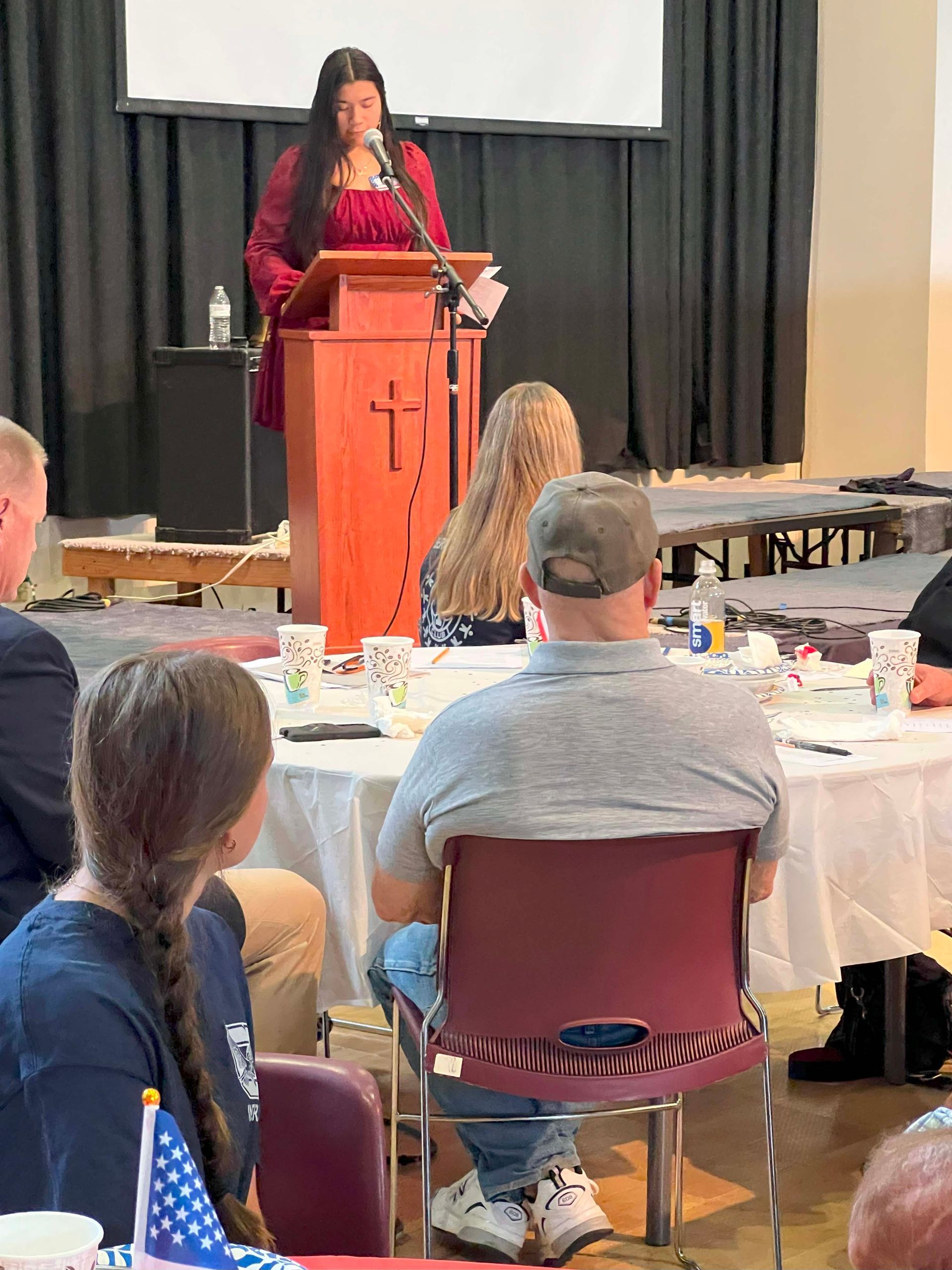 Woman in red speaking at a podium to an audience in a meeting hall