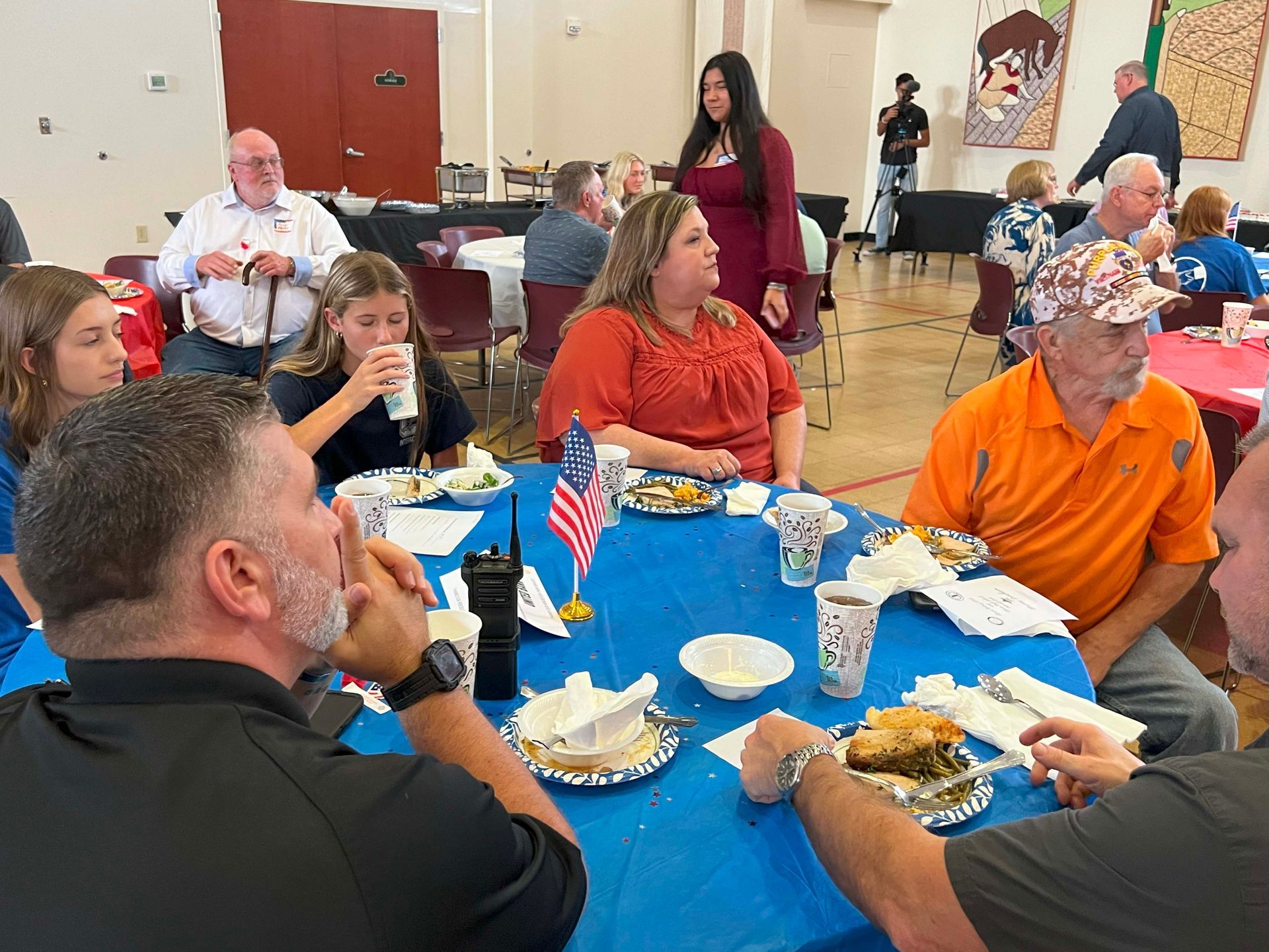People seated around a blue tablecloth at a social gathering, eating and chatting in a community room.