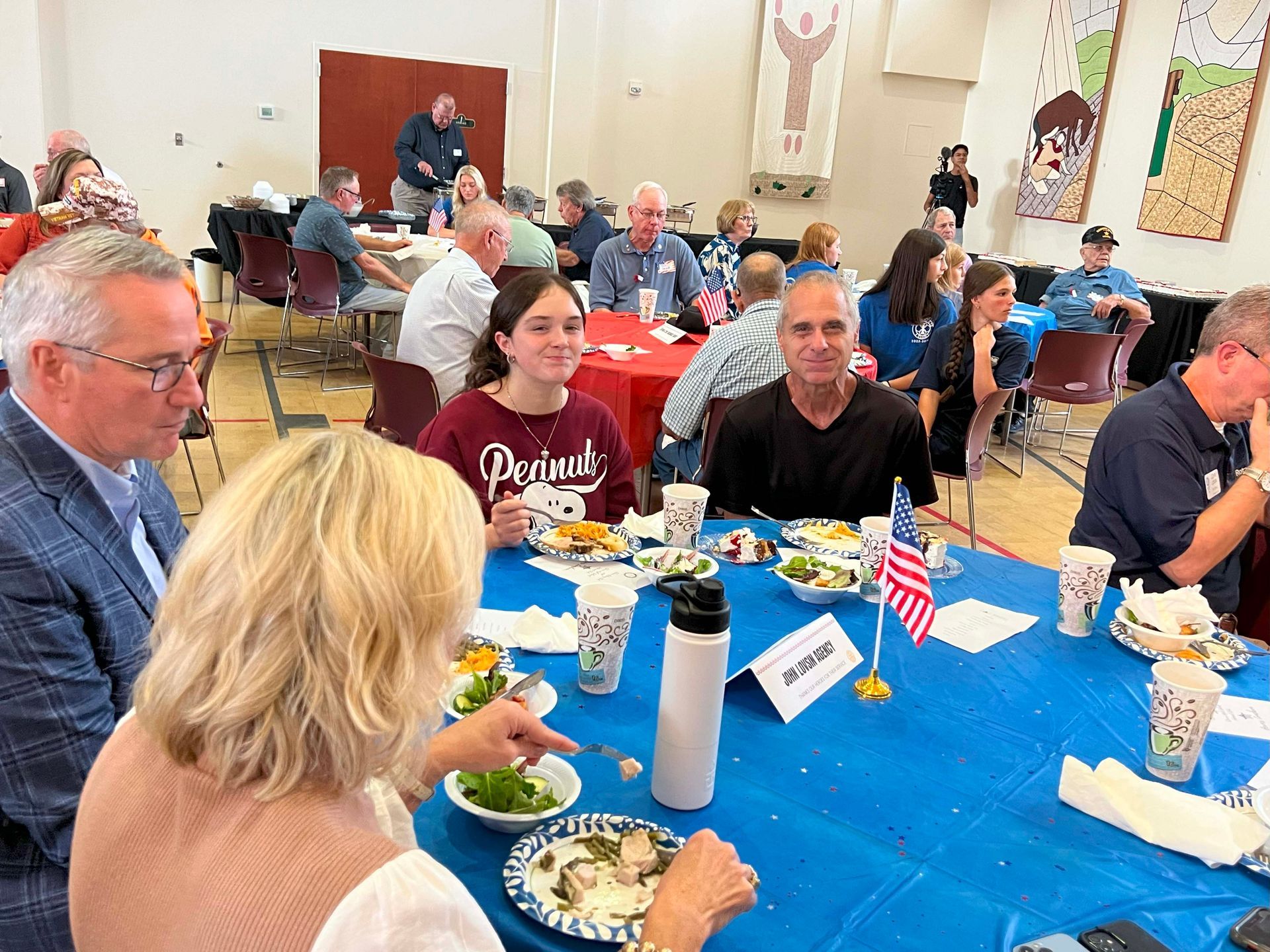 People seated at tables with food and drinks in a bright community hall, chatting during a meal.