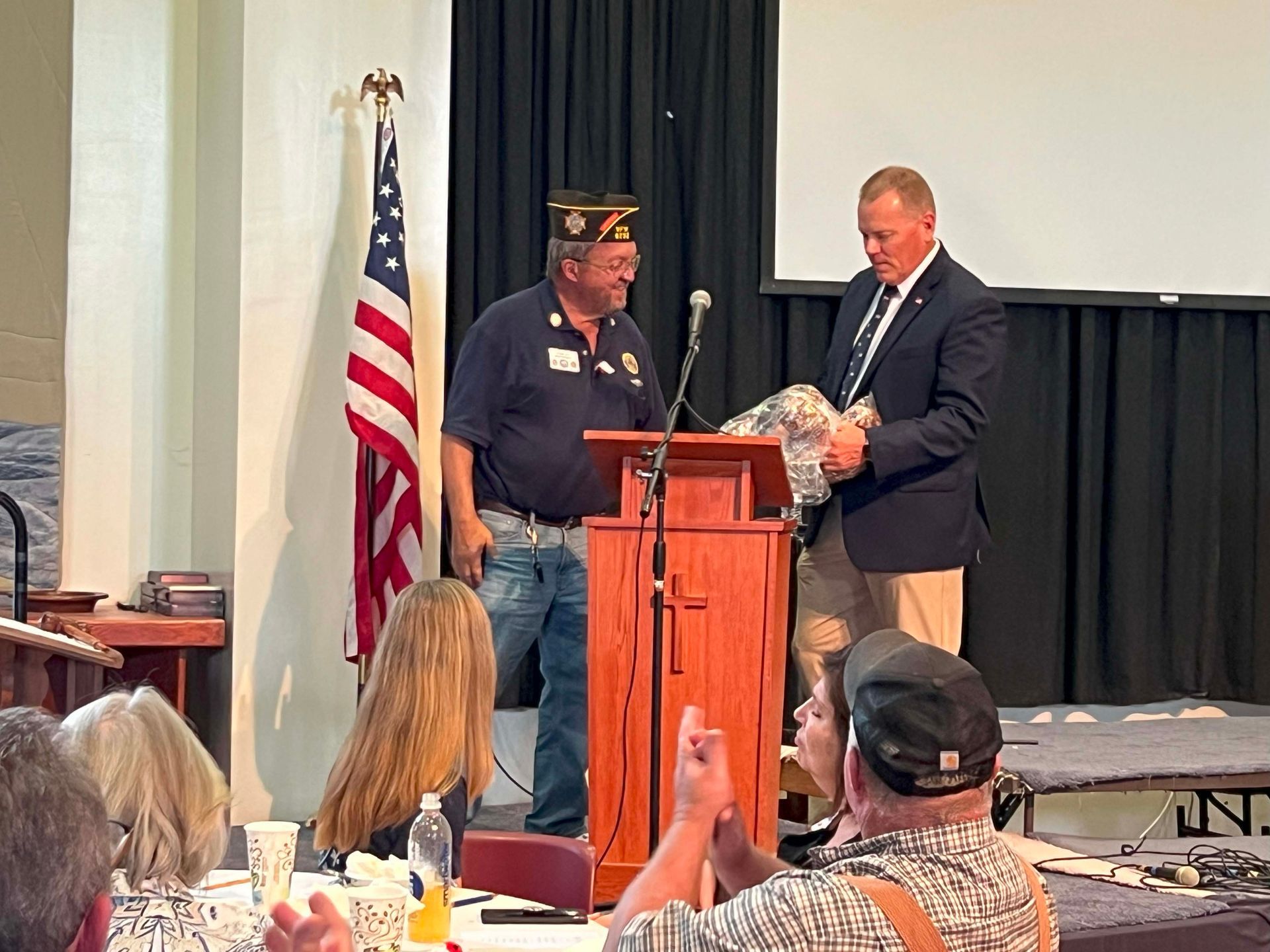 Man presents a gift to a veteran at a podium during a ceremony, with an audience and U.S. flag nearby.