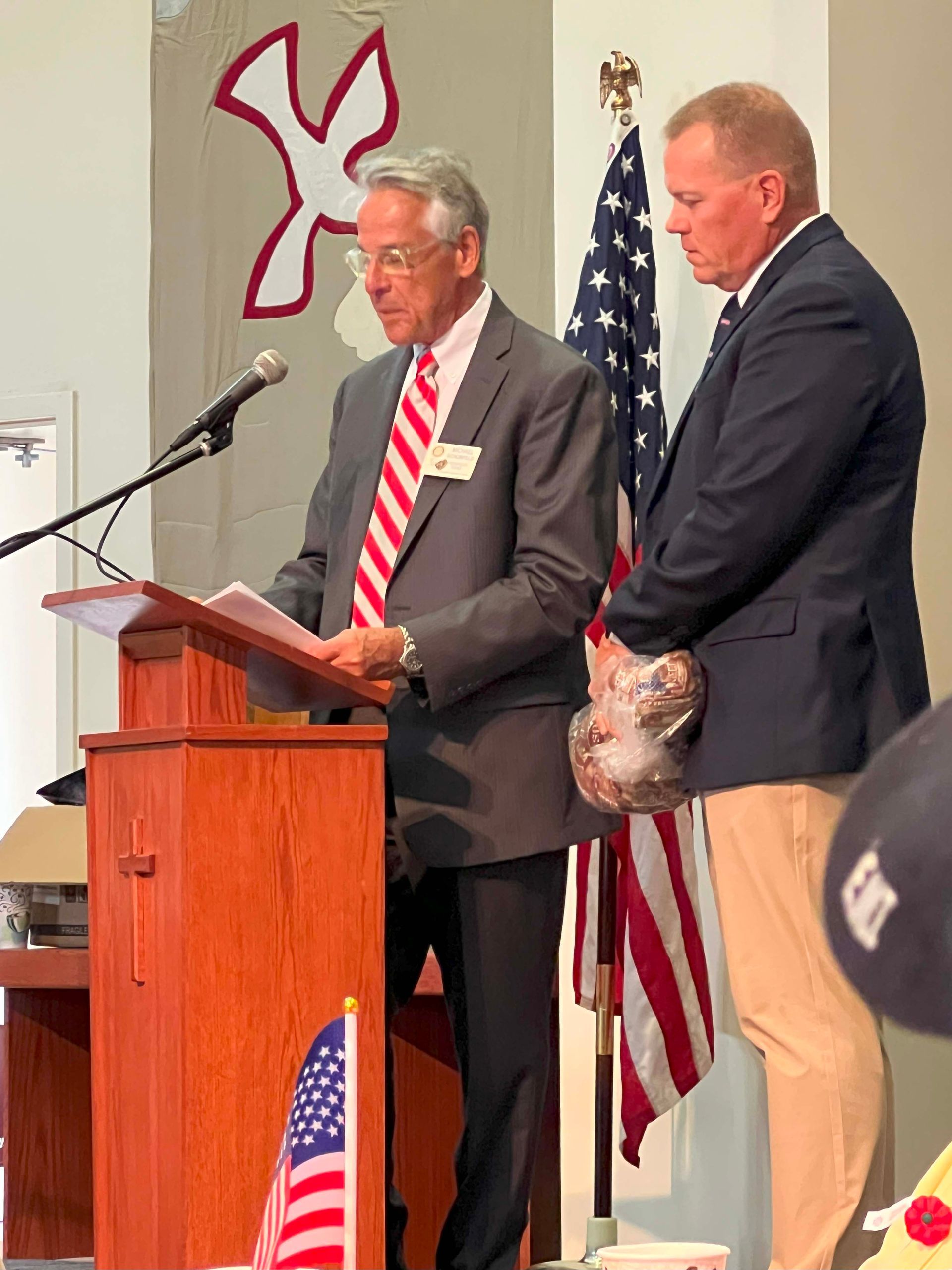 Two men standing at a podium during a ceremony, with U.S. flags in the background.