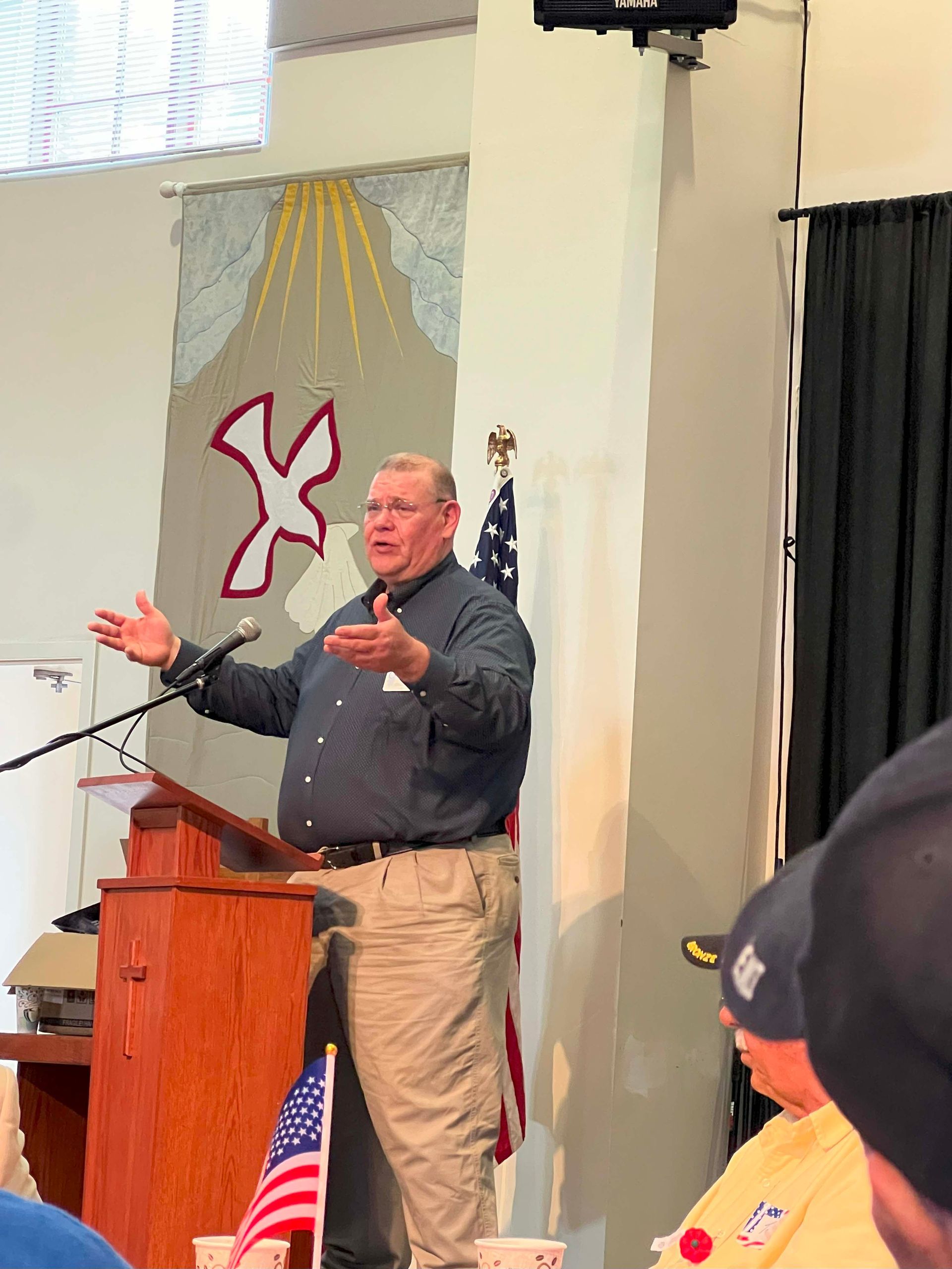 Man speaking at a podium with arms raised in a church or hall, American flag visible nearby.