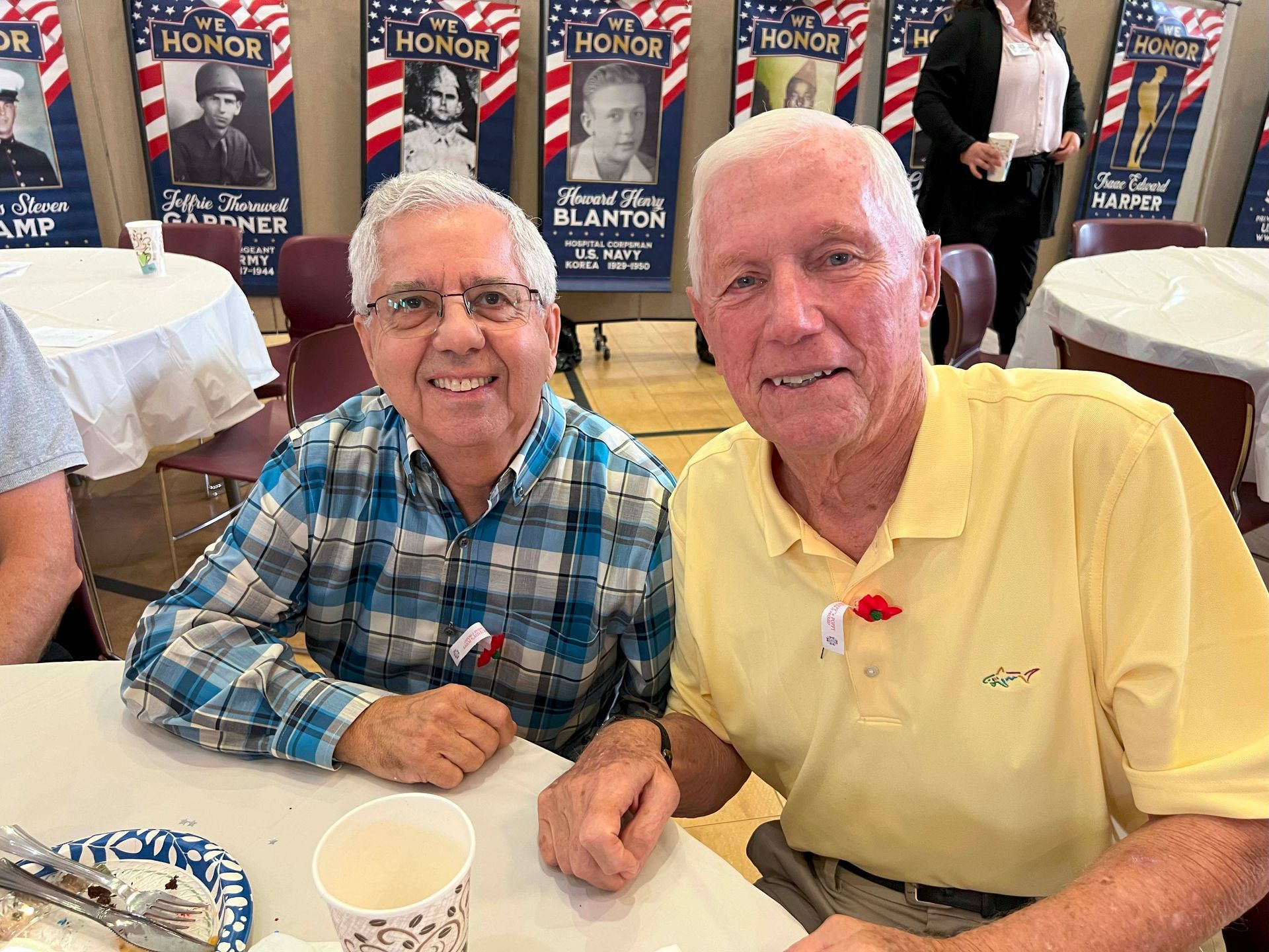 Two men smiling at a banquet table, one in a blue plaid shirt and one in a yellow polo.