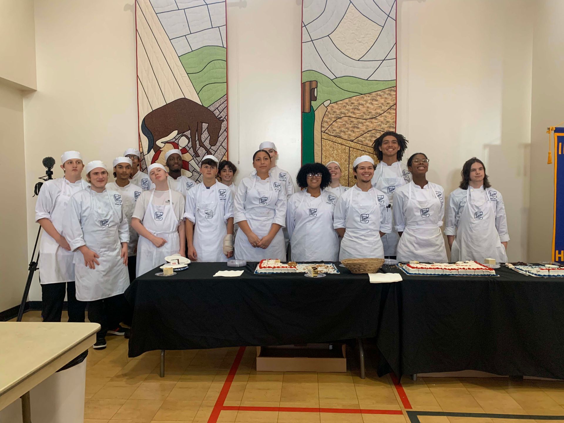 Group of chefs in white uniforms standing behind a table of dishes in a dining hall