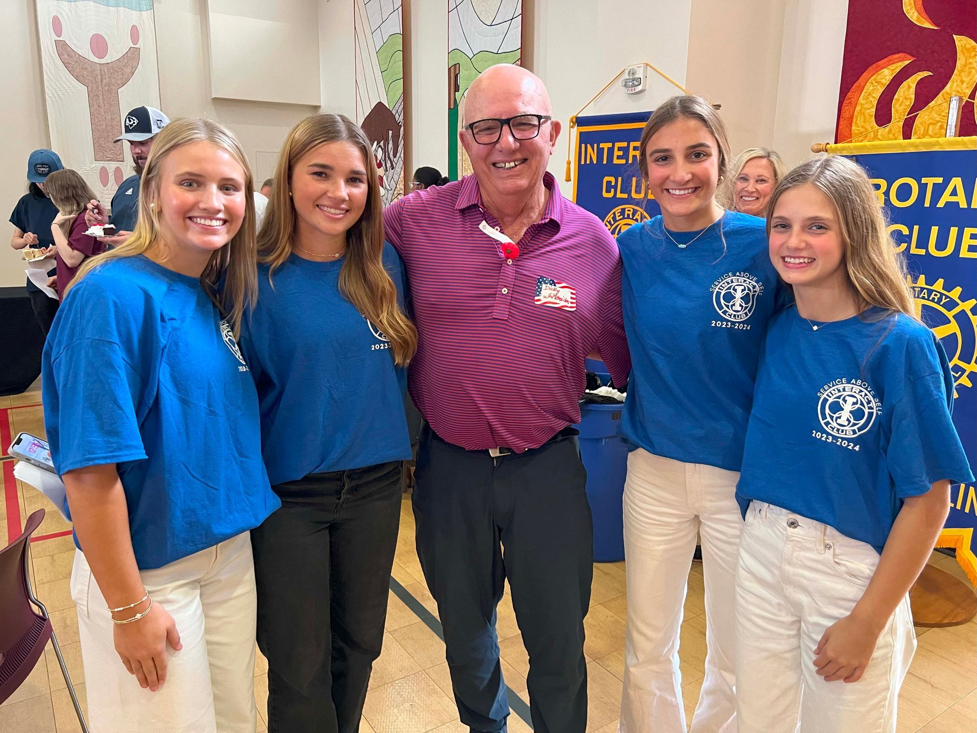 Five people smiling in blue shirts with a man in a maroon polo at an indoor event booth.