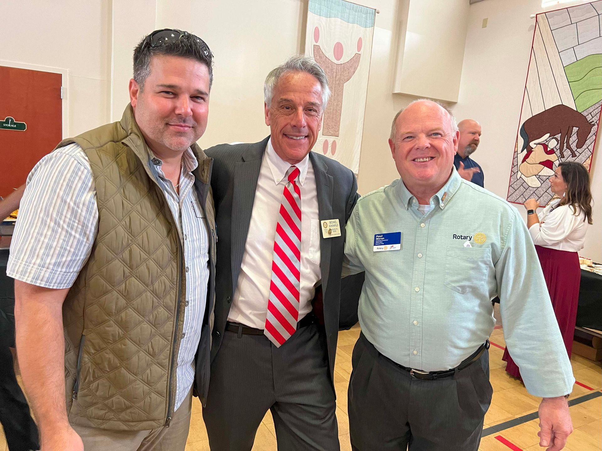 Three men smiling at an indoor event, standing together in front of posters and a table.