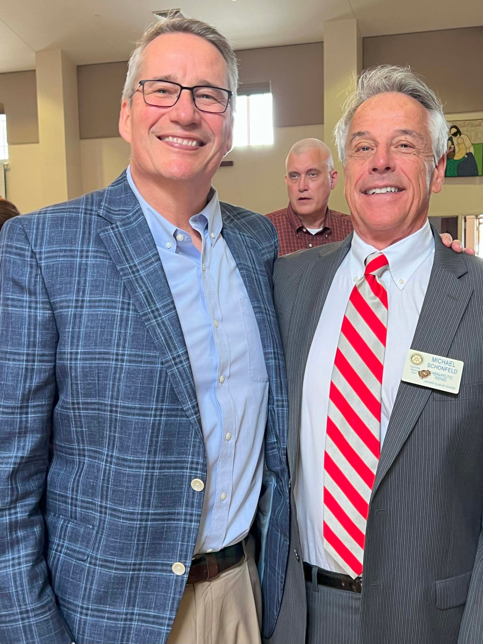 Two men smiling in suits at an indoor event, with a third man standing behind them.