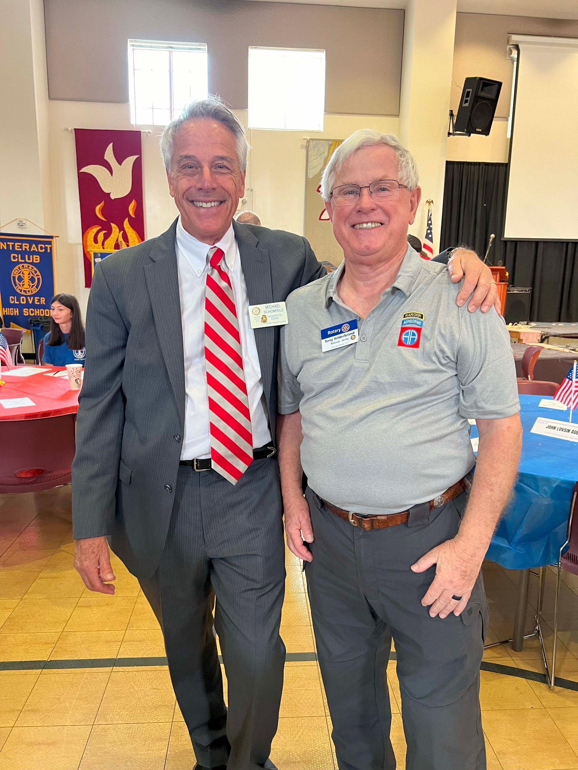 Two men smiling and posing together at an indoor event, one in a suit and tie, the other in a gray polo.