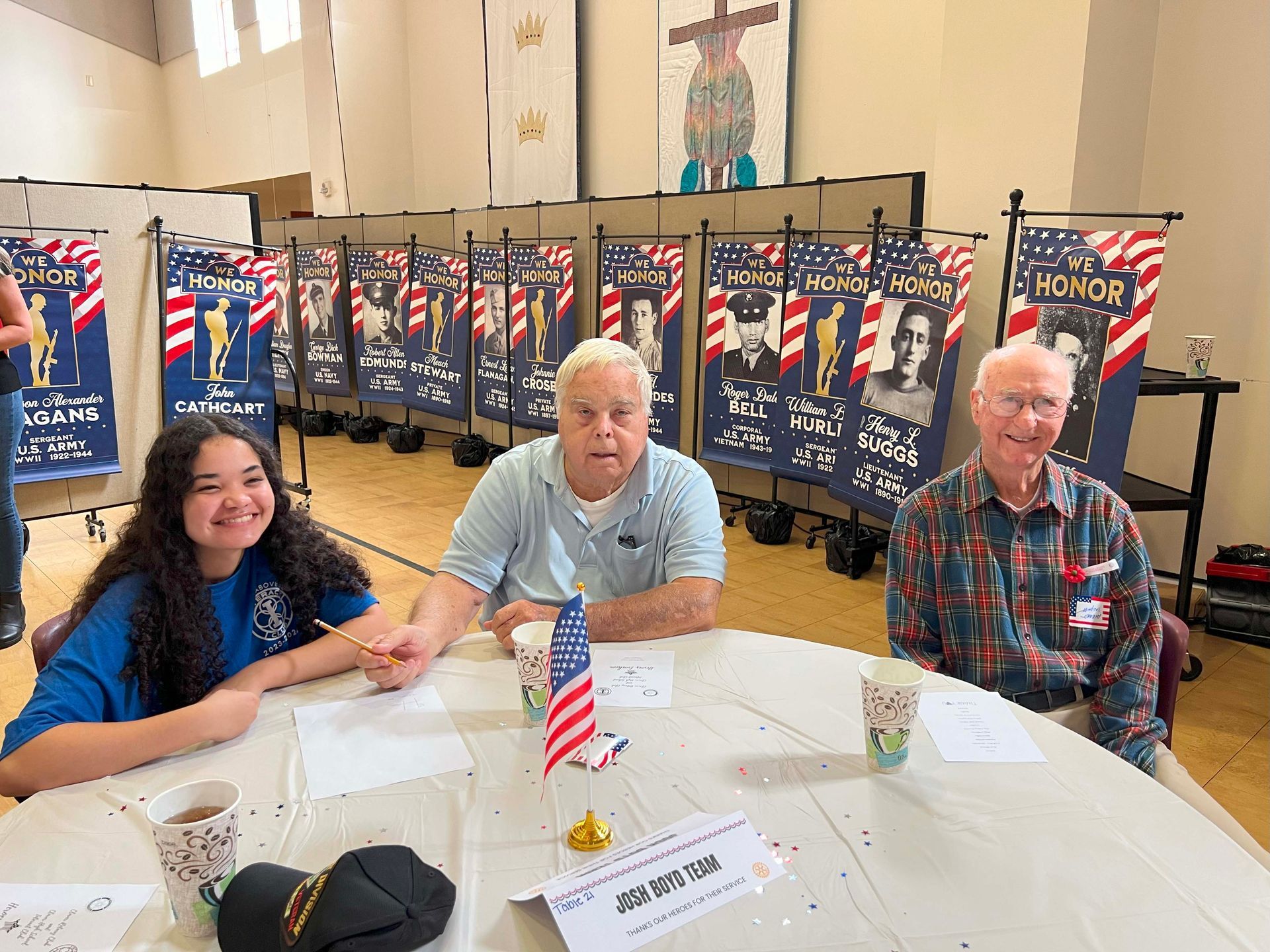 Three people seated at a table in a display hall, with patriotic banners and small flags behind them.