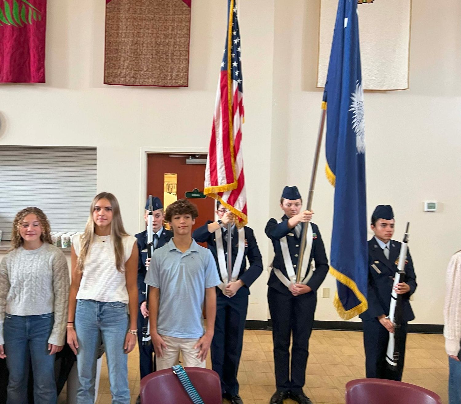Students standing indoors in a line with flags, including the U.S. flag, at a formal ceremony