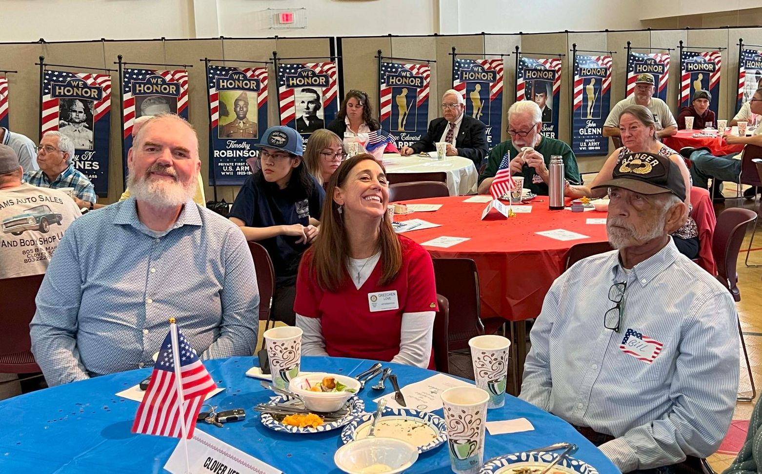 People seated at red-tablecloth tables in a hall decorated with small American flags, smiling and dining.