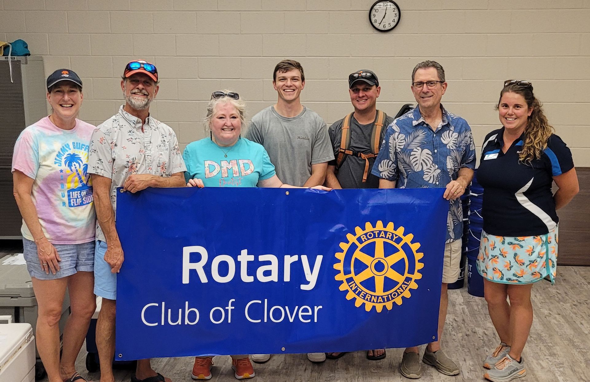Seven people holding a blue Rotary Club of Clover banner in a meeting room