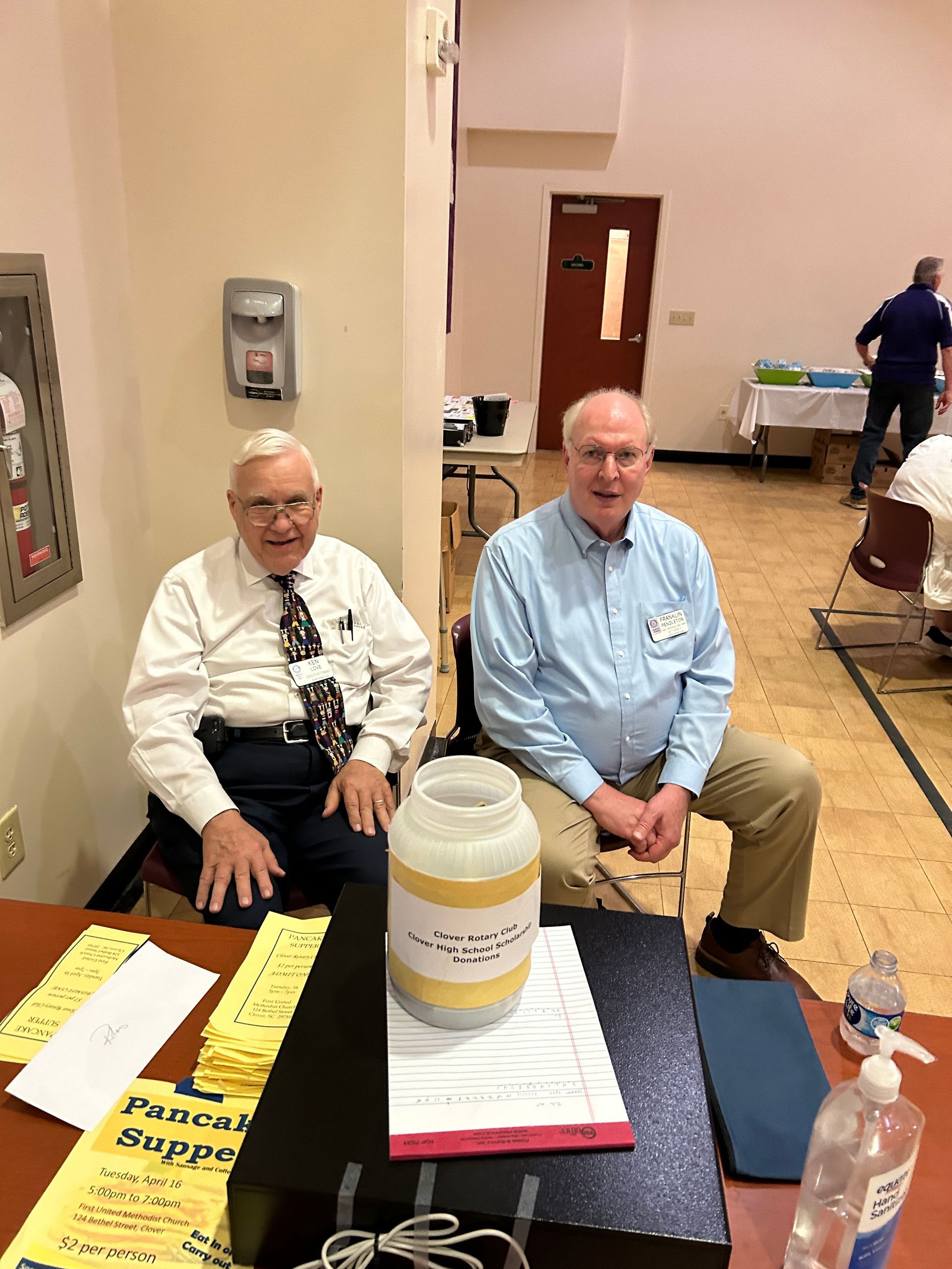 Two men seated at a table with papers and a donation jar in a gym-like indoor setting