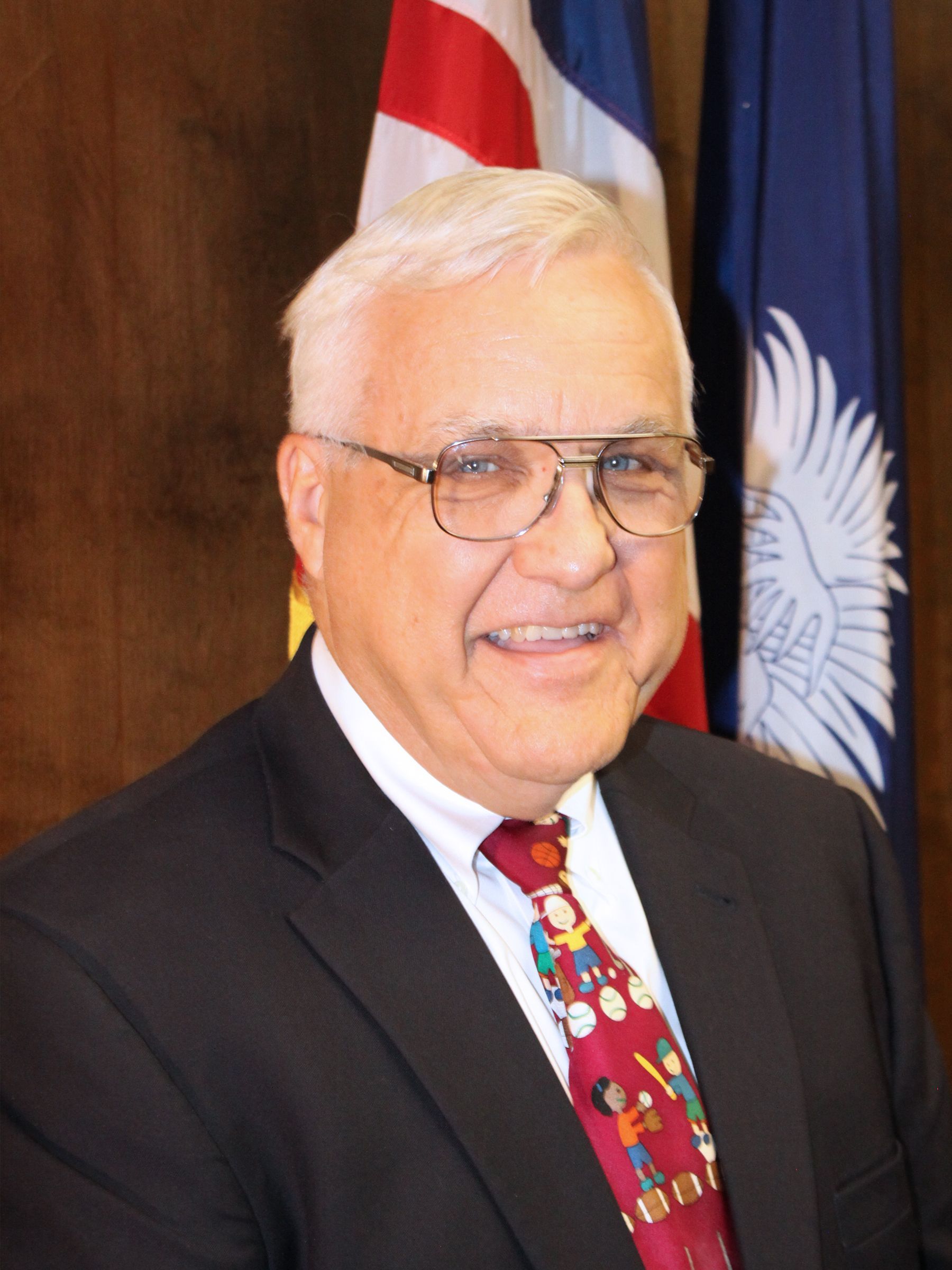Man in a dark suit and patterned tie smiling in front of flags and a wood-paneled wall