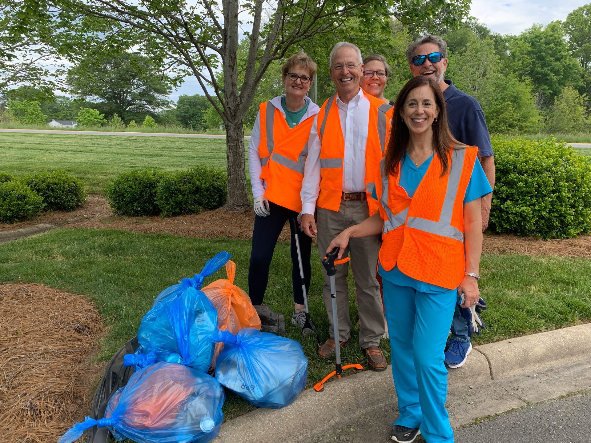 Volunteers in orange safety vests stand by trash bags during a roadside cleanup in a park.