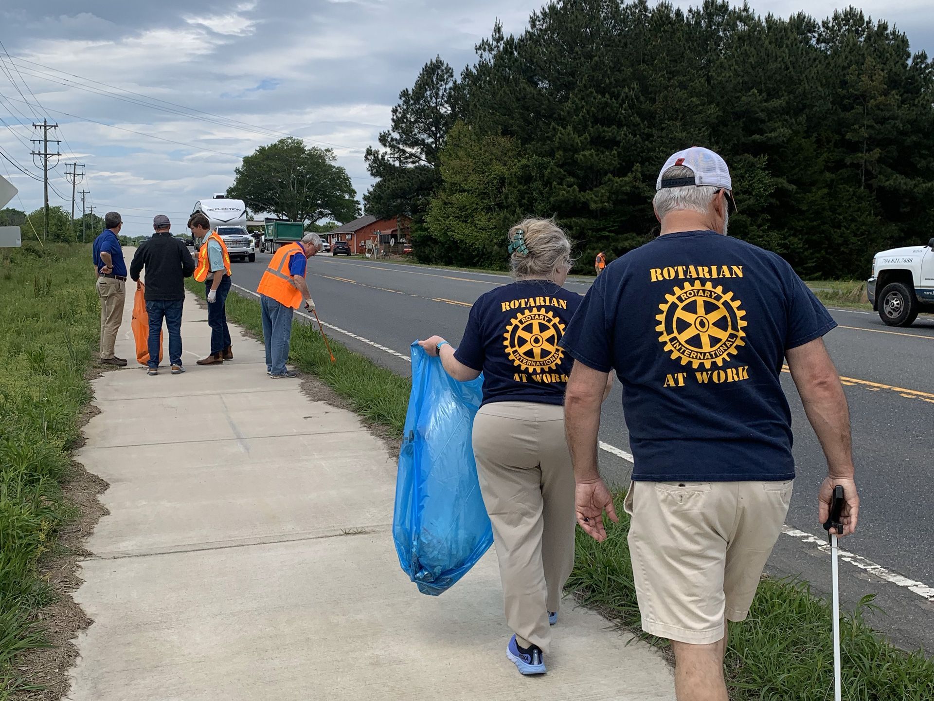 Volunteers in navy shirts clean roadside litter along a sidewalk and road edge, carrying a blue trash bag.
