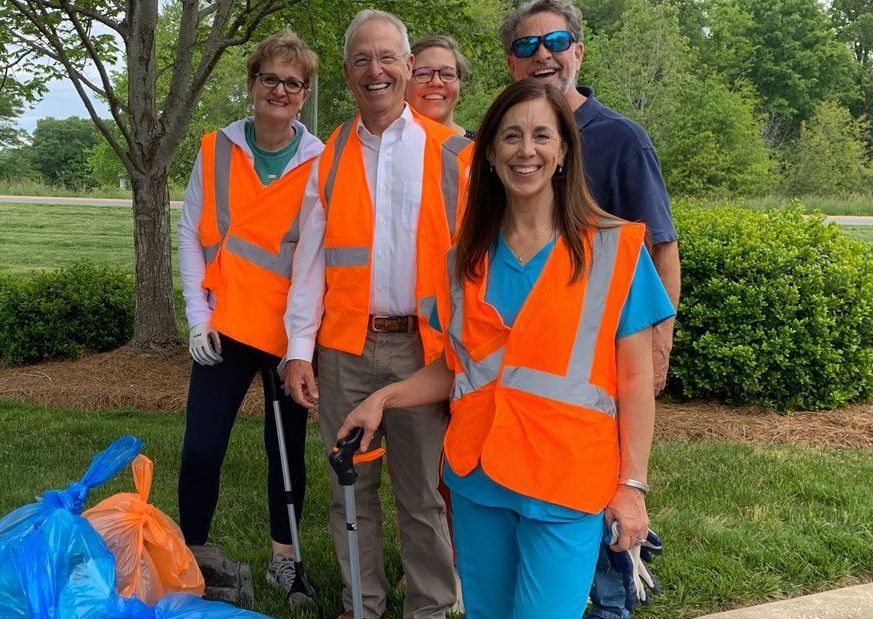 Four volunteers in orange safety vests stand outdoors by a grassy path and trees, smiling at the camera.