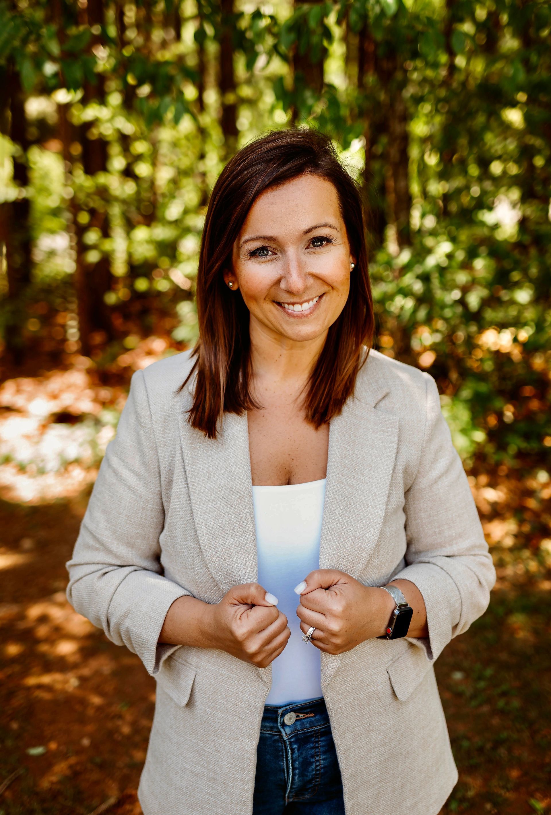 Woman in beige blazer smiling outdoors in a wooded setting.