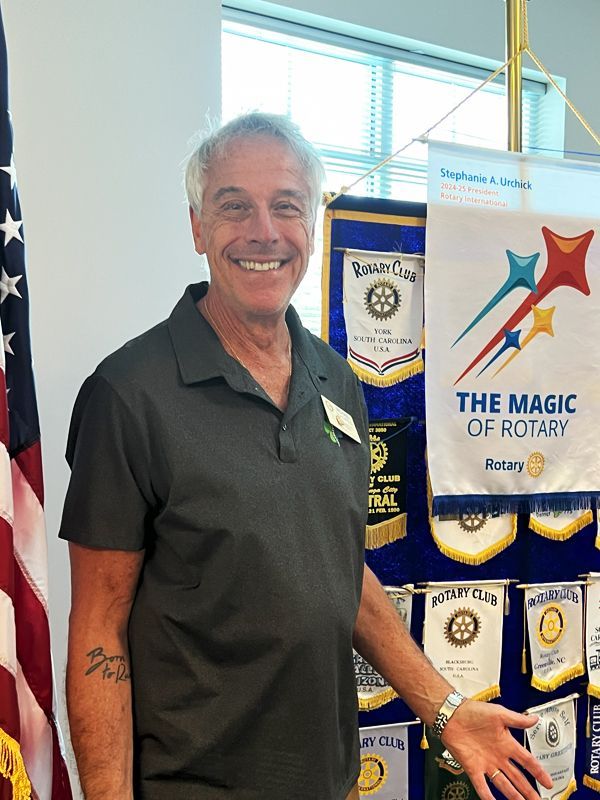 Smiling man in black polo stands beside a Rotary “The Magic of Rotary” banner and American flag