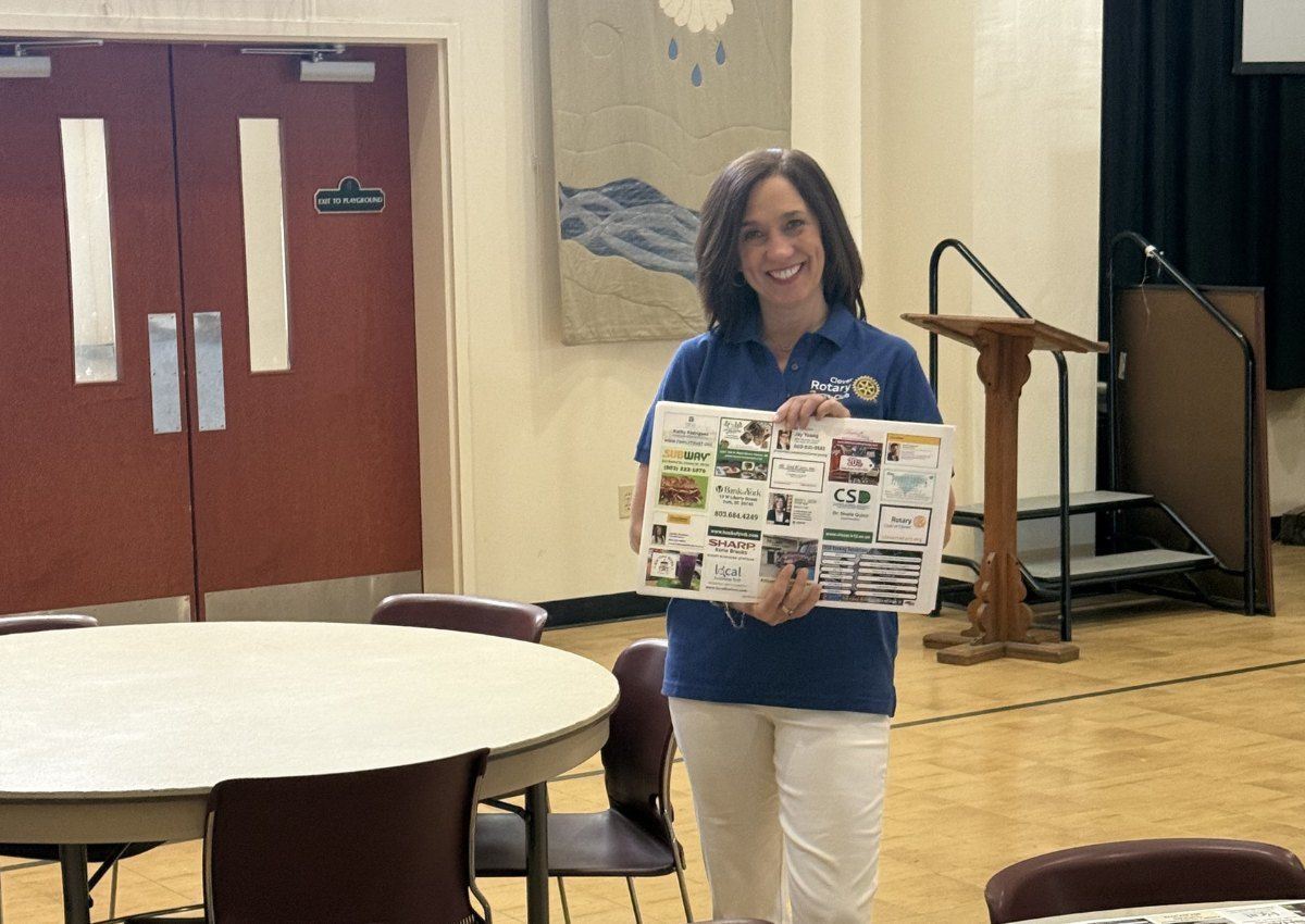 Woman in blue shirt holds a poster board in a community hall near tables and a podium.