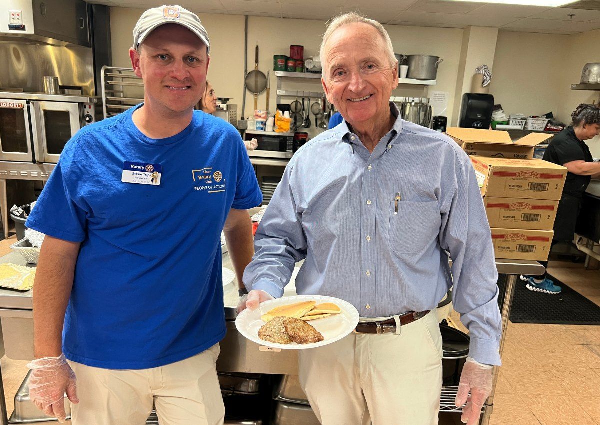 Two men in a kitchen hold a plate of food, one in a blue shirt and the other in a dress shirt.