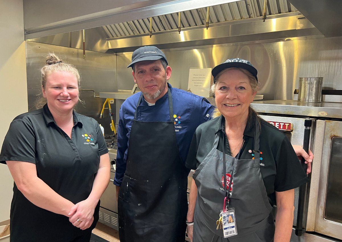 Three kitchen staff in black and blue uniforms standing in a stainless-steel commercial kitchen