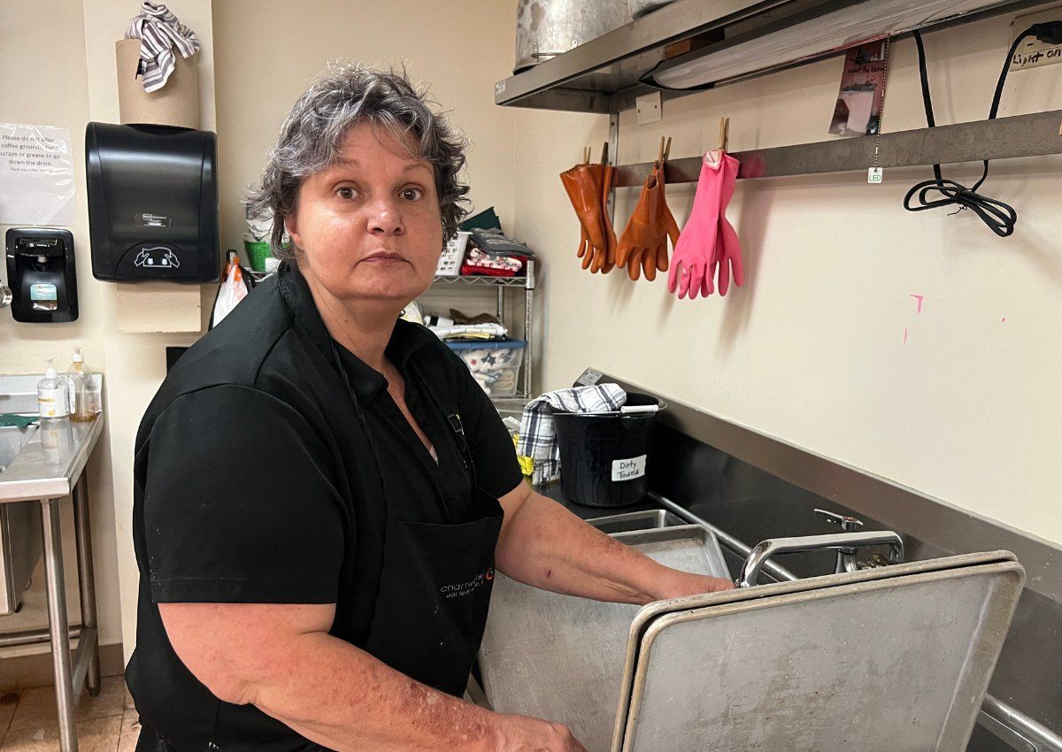 Woman standing at a stainless-steel sink in a commercial kitchen, wearing a black shirt and apron.