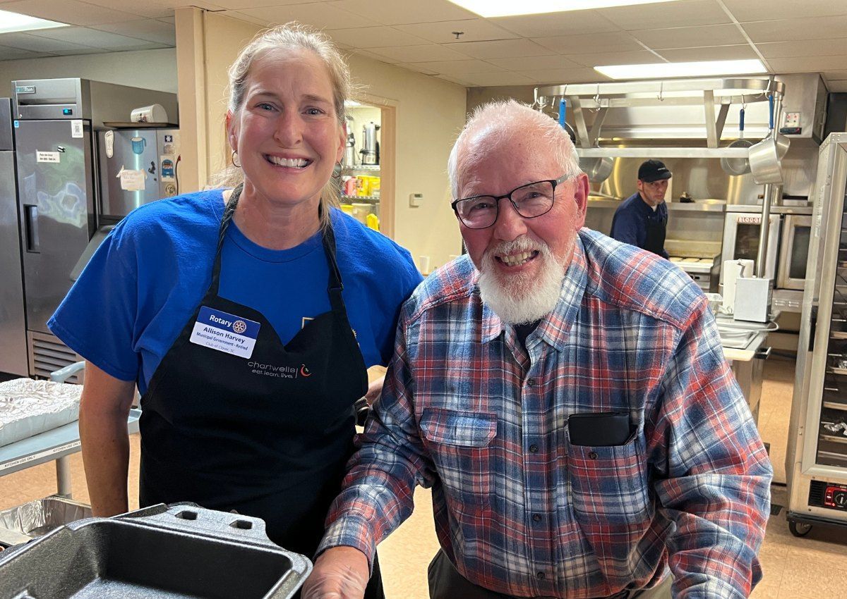 Two smiling people in a commercial kitchen, wearing aprons and standing by prep counters.
