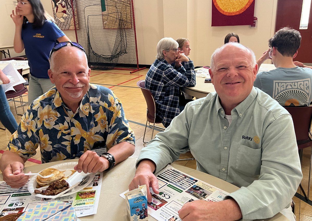 Two men seated at a cafeteria table, smiling over lunch with newspapers on the table