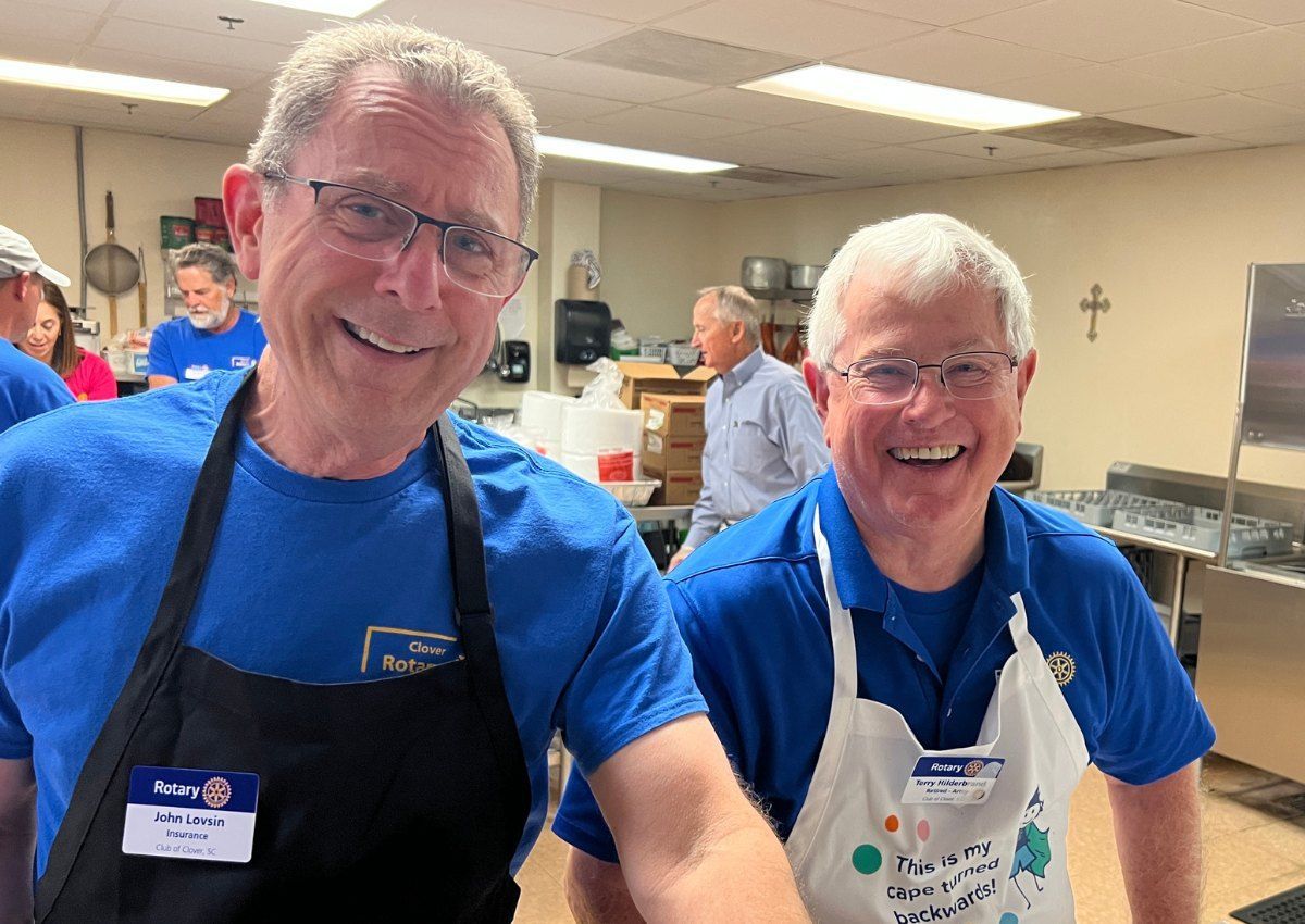 Two smiling volunteers in blue shirts and aprons in a kitchen serving area