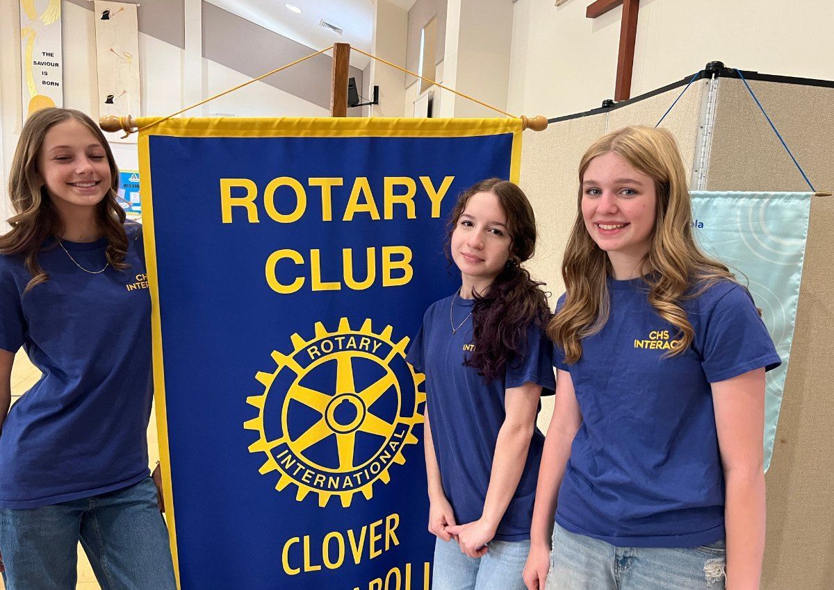 Three people in blue shirts stand beside a Rotary Club Clover banner.