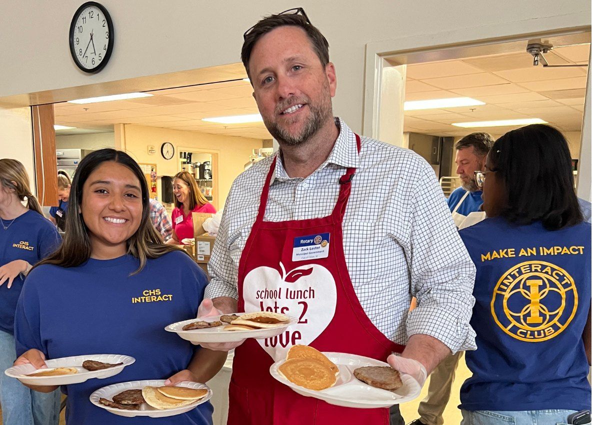 Two people in aprons hold plates of food in a bright indoor room with others in the background.
