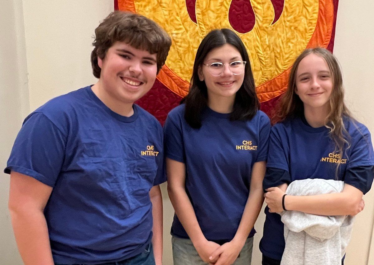 Three people in blue T-shirts stand smiling in front of a large red and gold mural.