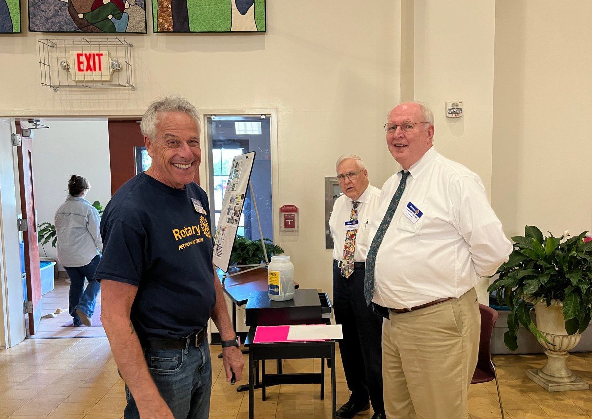 Three men stand smiling in a bright lobby beside a small table and potted plant.