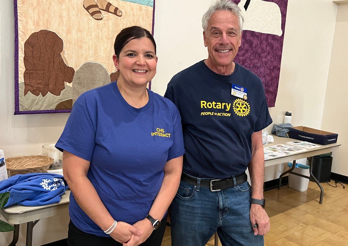 Two people smiling indoors beside a table, wearing Rotary shirts and standing in front of artwork.