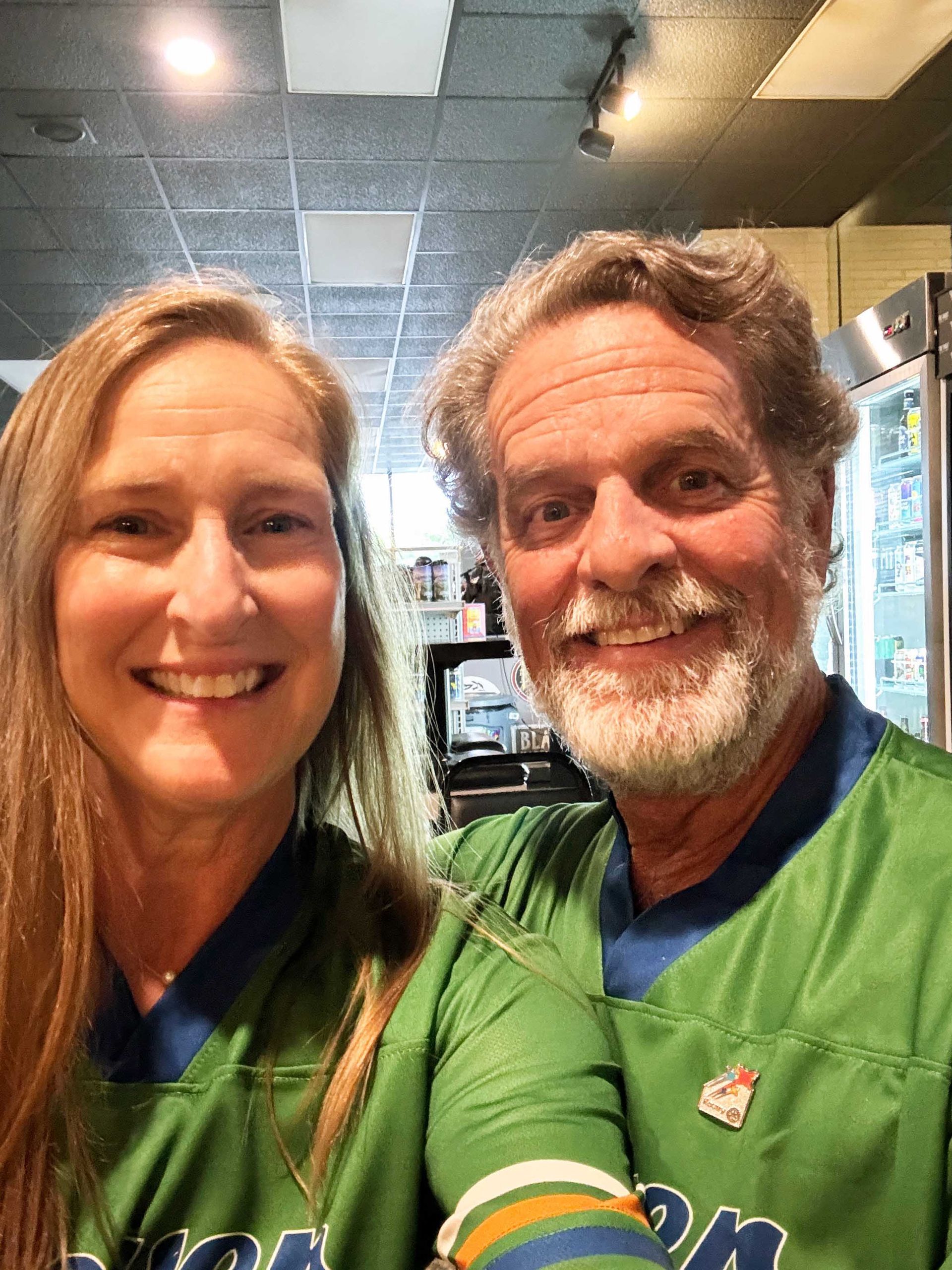 Smiling pair in green shirts taking a selfie indoors at a shop or café