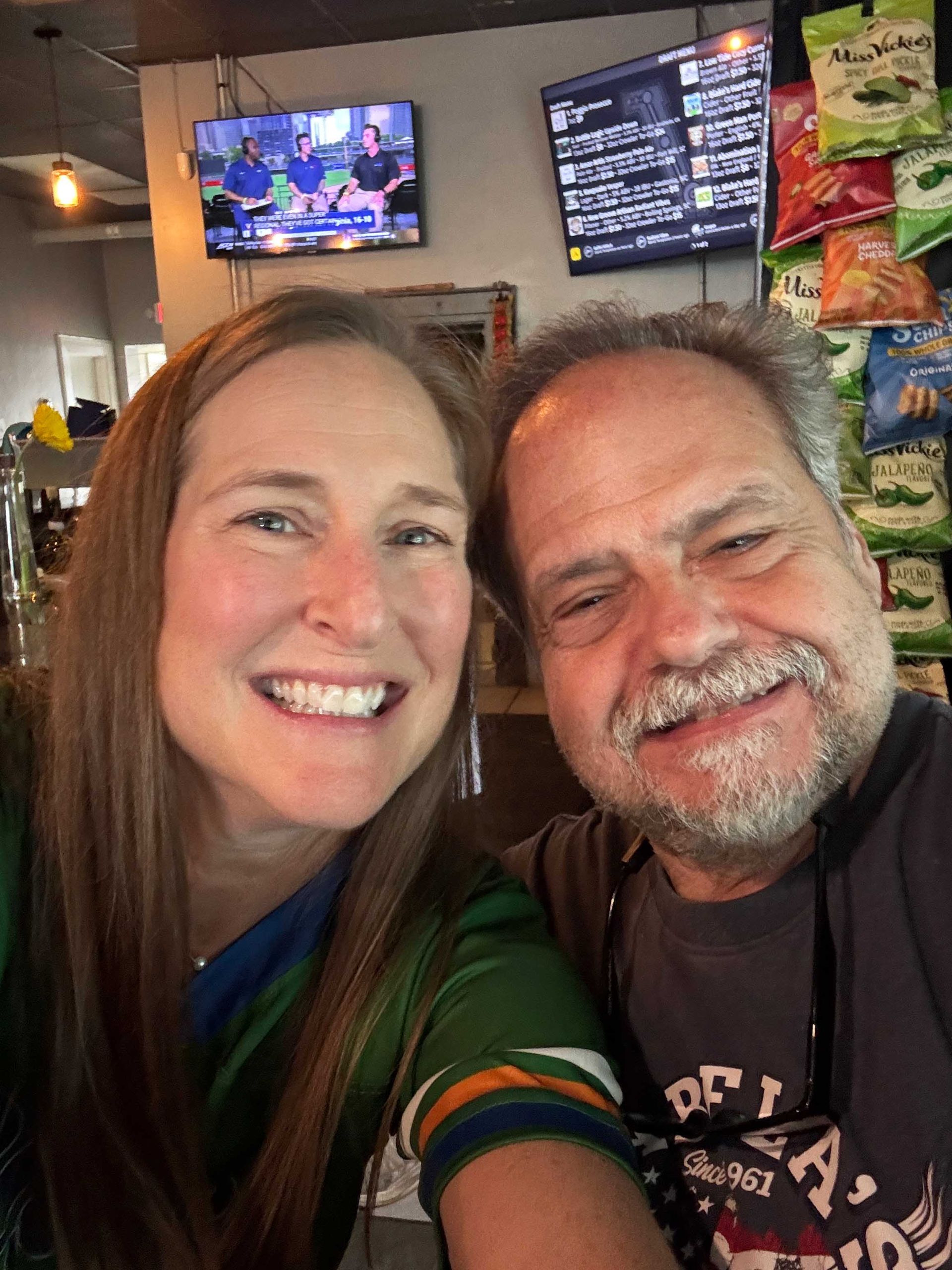 Smiling couple taking a selfie in a lively bar, with TVs and snack displays in the background.