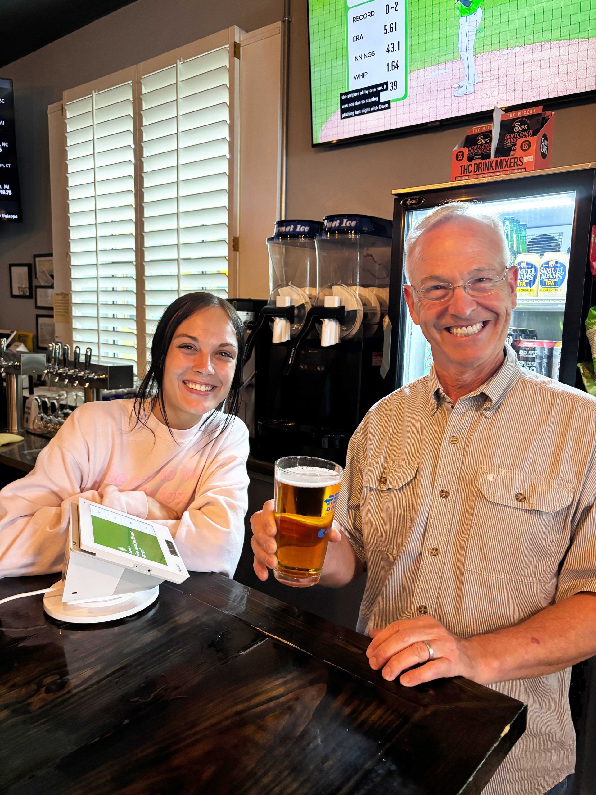 Two people smiling at a bar, holding drinks, with a TV and window shutters behind them.
