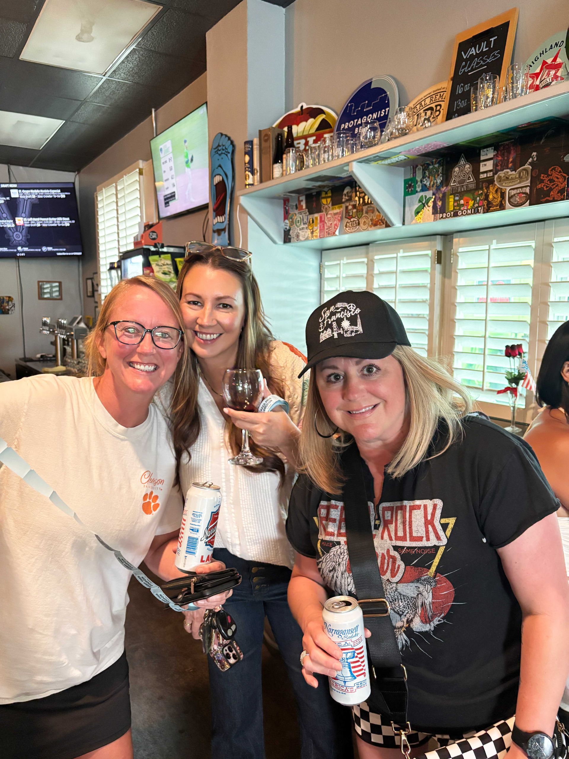 Three smiling people posing together in a shop, one holding a drink, with colorful shelves behind them