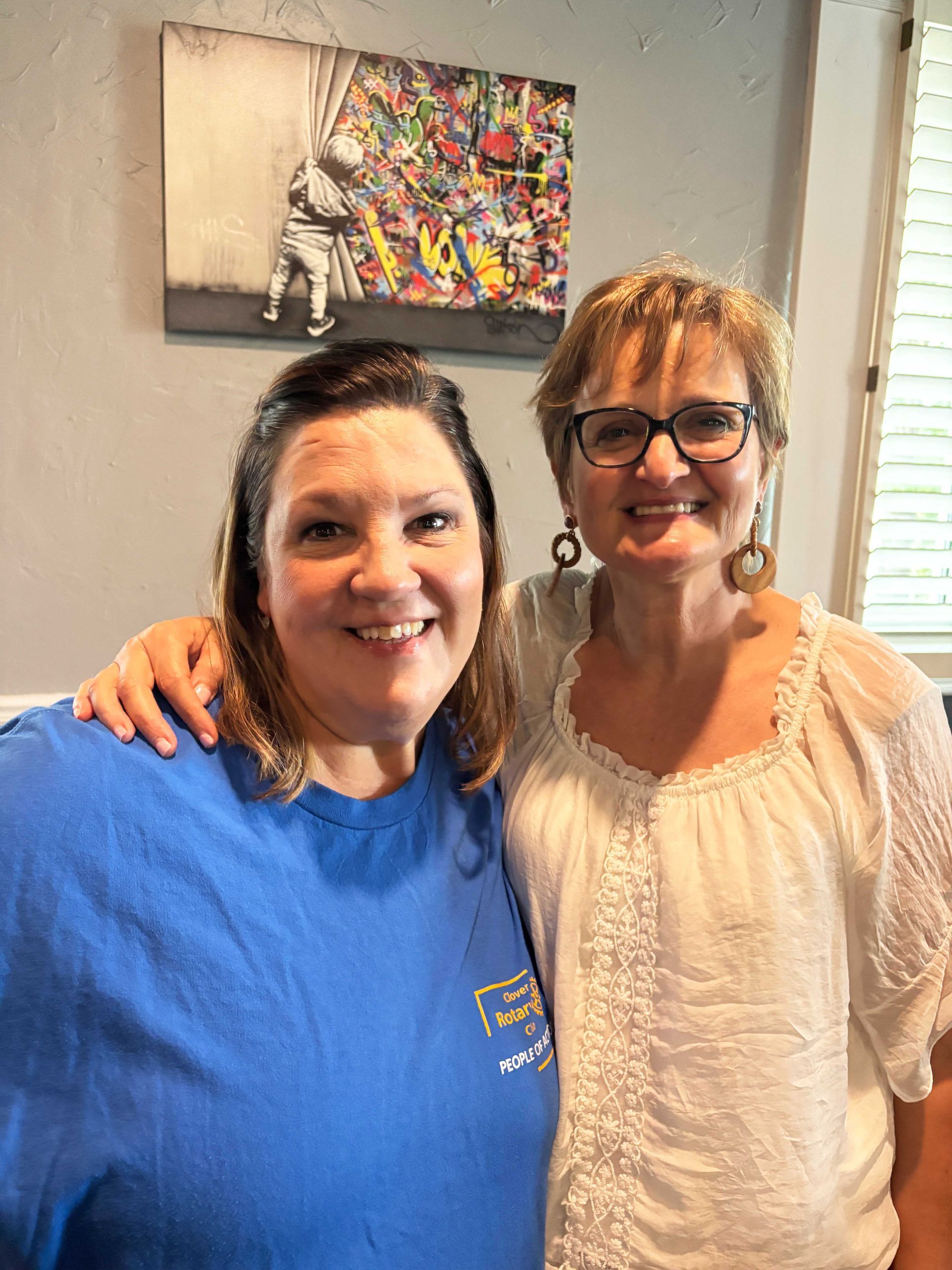 Two smiling people standing indoors, one in a blue shirt and one in a white blouse, with wall art behind them