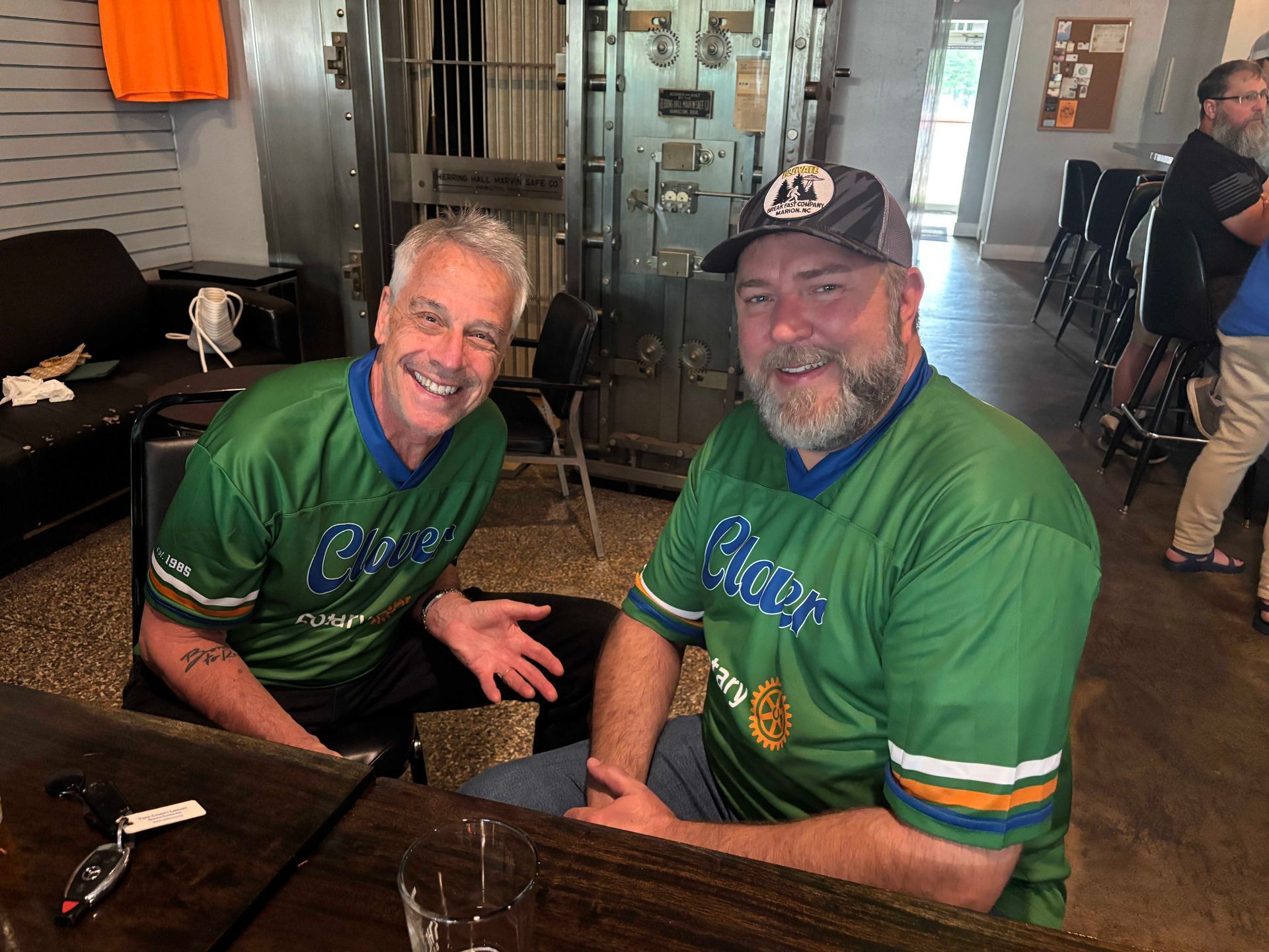 Two smiling men in green jerseys sit at a table indoors, posing together.