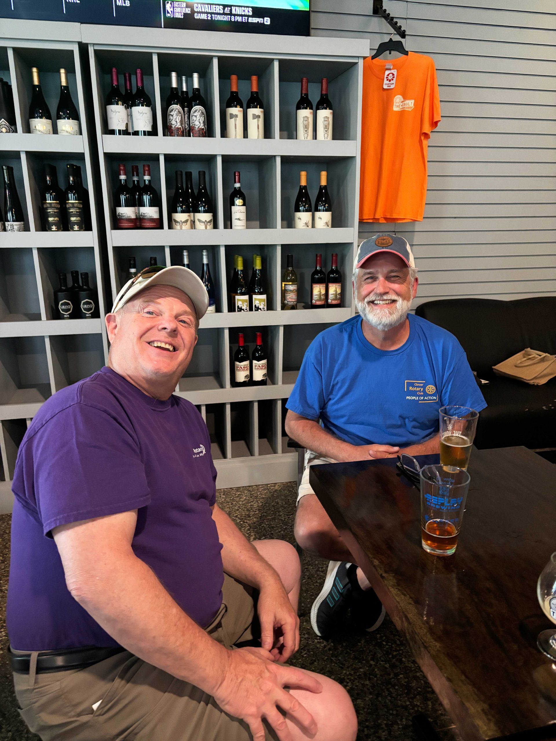 Two smiling people seated at a wooden table in a bar, with shelves of wine bottles behind them.