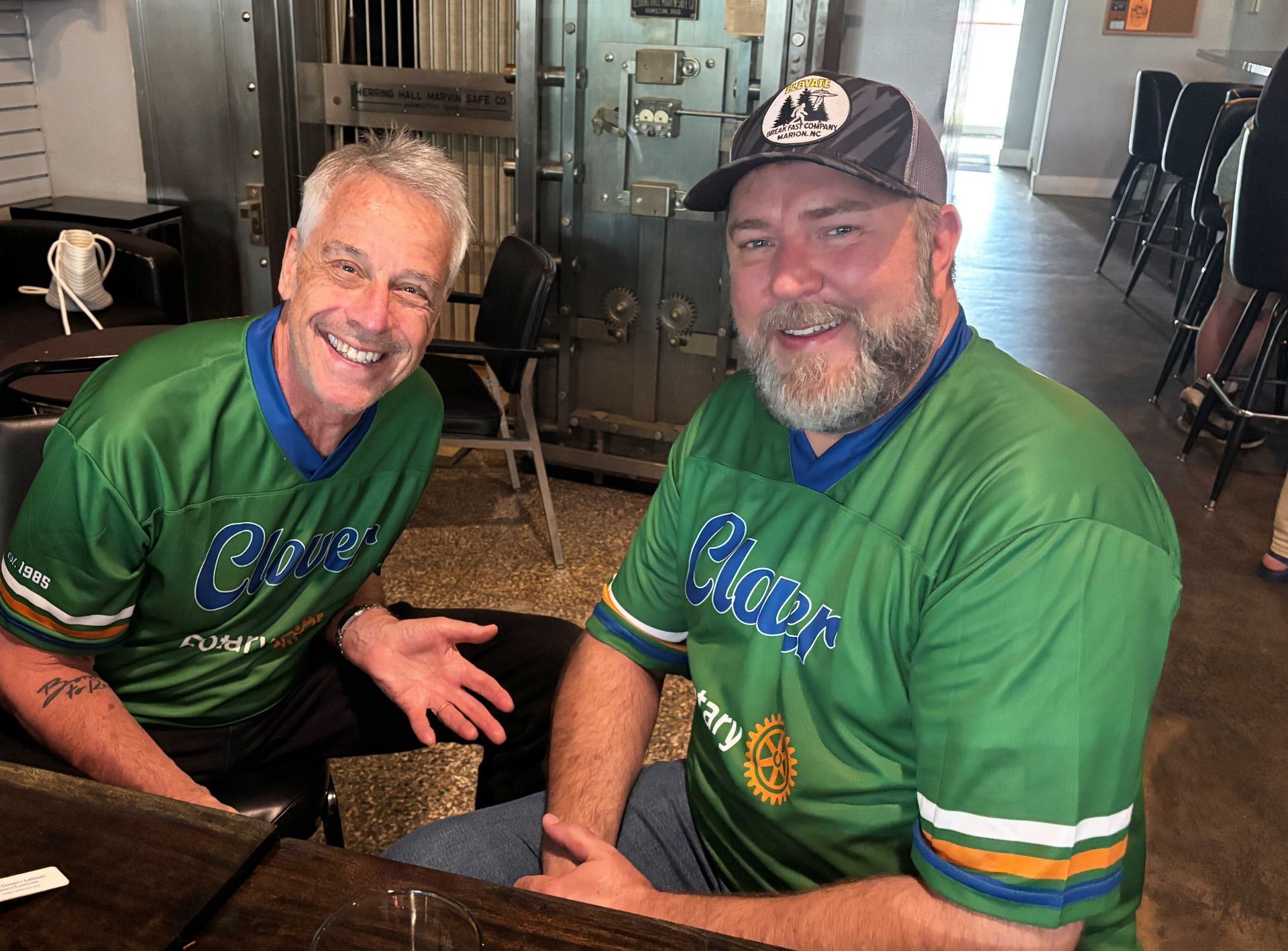 Two smiling men in green jerseys seated indoors at a table, wearing baseball caps.