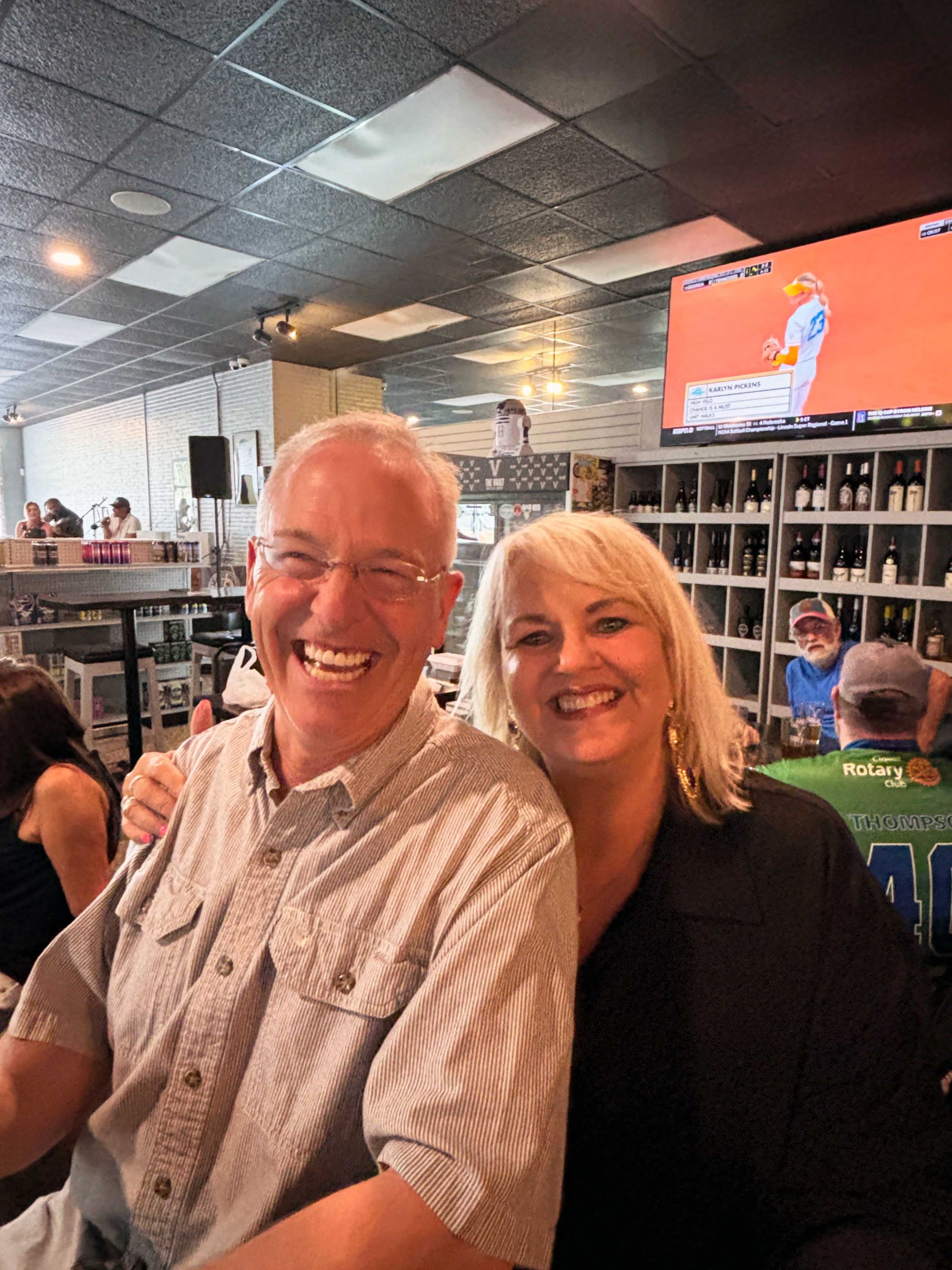 Two smiling people seated in a lively bar, with shelves and a TV screen in the background.