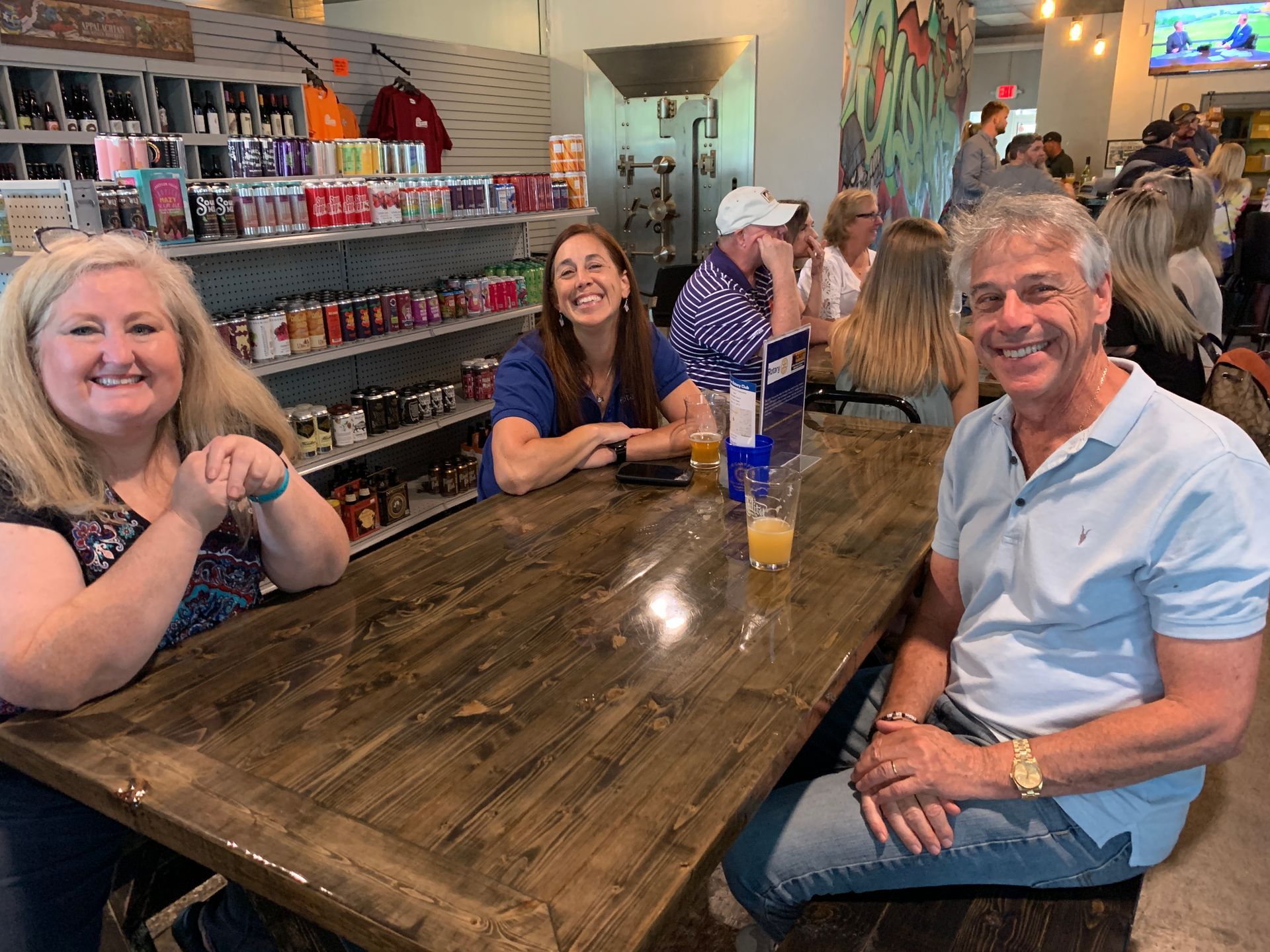 Three people smiling at a wooden table in a lively bar or café, with drinks and shelves in the background