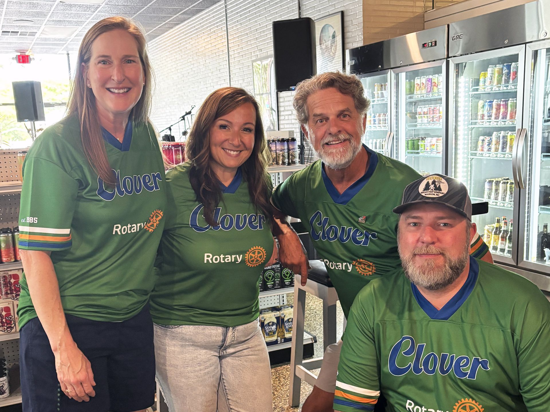 Four people in green Clover Rotary shirts smiling in a refrigerated store aisle