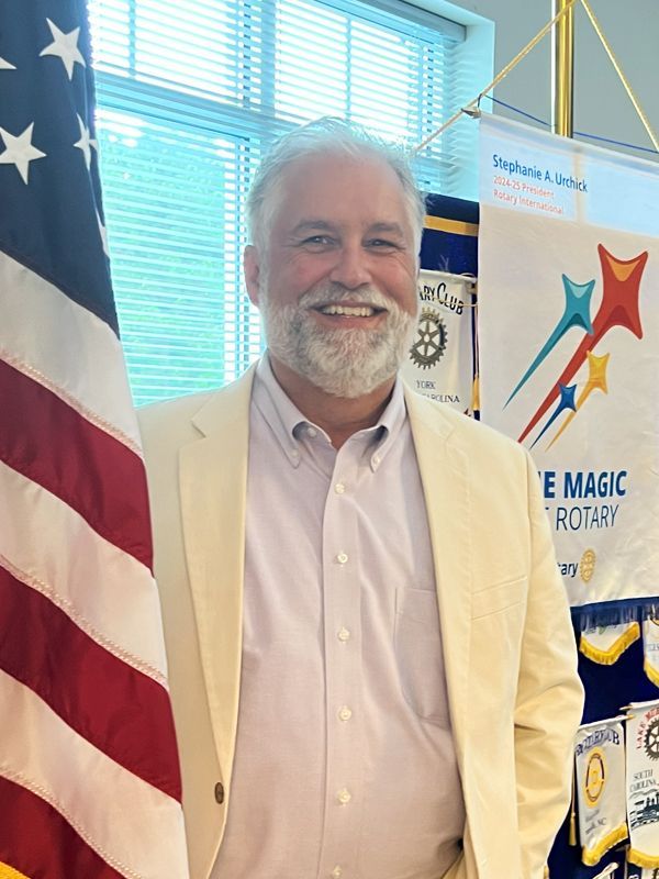 Smiling man in a cream jacket standing beside an American flag at a Rotary Club event