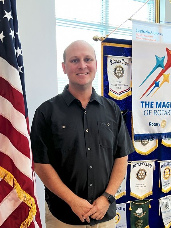 Man in black shirt standing beside U.S. flag and award banners in an office.