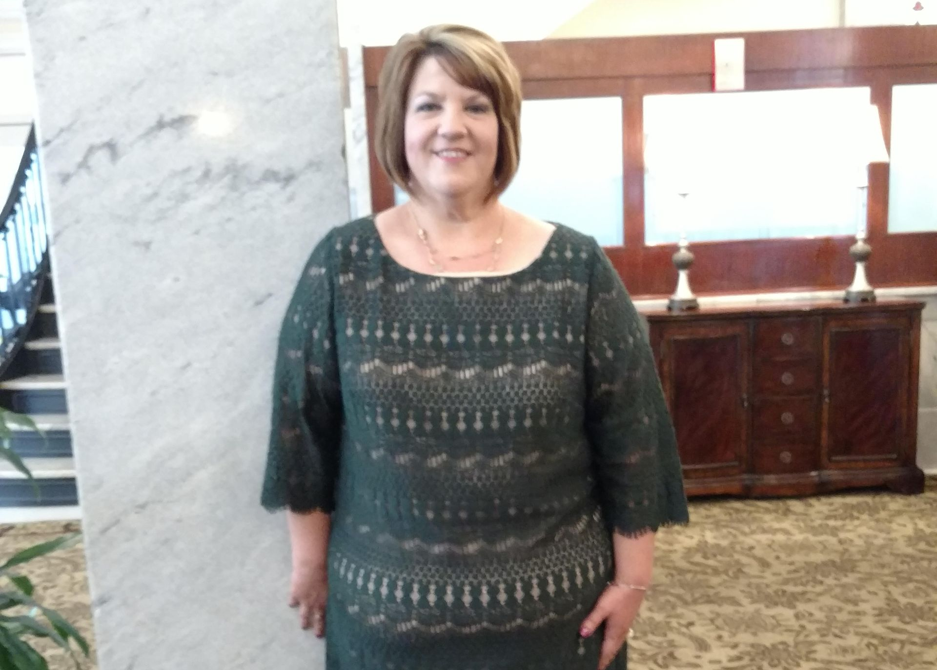 Woman in a dark green dress standing in a lobby beside a marble column and wooden cabinet.