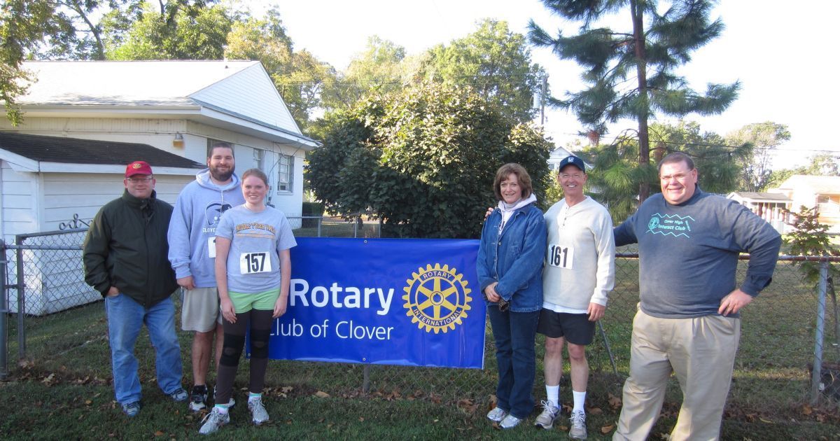 Five people standing by a Rotary Club of Clover banner outdoors near a house and trees.